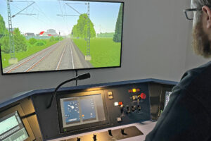Ein Mann sitzt vor einem Bedienpult eines Zuges und vor einem Bildschirm, der eine Bahnstrecke zeigt.